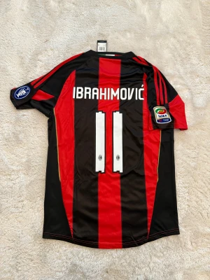 AC Milan Ibrahimović matchtröja - Säljer en officiell AC Milan fotbollströja från Adidas med Ibrahimović och nummer 11 på ryggen. Tröjan är kortärmad, har klassiska röda och svarta vertikala ränder, klubbmärke på bröstet och Serie A-patch på ärmen. Materialet är lätt och andas, perfekt för fotboll eller samling.