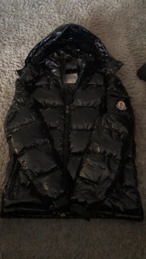 Svart Moncler maya  - Säljer en svart, glansig pufferjacka från Moncler med huva och tydlig logga på ärmen. Jackan har dragkedja med Moncler-detalj och är quiltad för extra värme. Perfekt för kalla dagar och har en riktigt snygg streetstil. Hojta till om ni har någon fundering:) går även att sänka i pris! Vill få den såld så snabbt som möjligt.