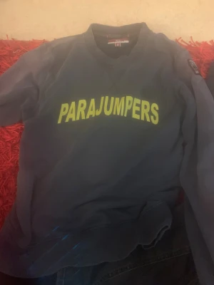 Mörkgrå fleece-tröja från Parajumpers - Säljer en mörkgrå fleece-tröja från Parajumpers med stor gul logga på bröstet och klassisk patch på ärmen. Tröjan har rund halsringning och ribbade muddar. Perfekt för kyliga dagar och enkel att matcha med jeans eller joggers. Tröjan är i okej skick och passar mig inte längre därav priset det låga priset köpt på nk för 999 kr på rea
