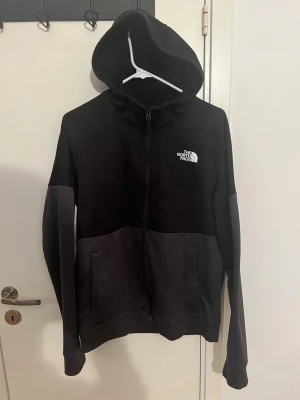 Svart hoodie från The North Face  - Svart hoodie från The North Face i storlek M. Tröjan har hel dragkedja, huva och två fickor framtill. Materialet är mjukt och känns som polyester, perfekt för chill eller träning. Snyggt vitt logotryck på bröstet och diskret ribbad struktur på tyget.