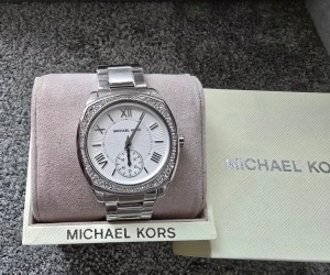 Michael Kors damklocka – helt ny, med box - Stilren Michael Kors-klocka i silver med metallänk och glittrande stenar runt boetten. Vit urtavla med romerska siffror och liten sekundvisare. Klockan är helt ny och oanvänd med skyddsplast kvar och levereras i originalask med kudde och manual. En elegant accessoar som passar alla tillfällen.