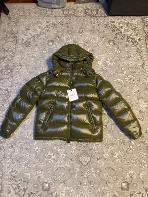 Olivgrön Moncler dunjacka med huva - Säljer en olivgrön dunjacka från Moncler med glansig finish, huva och två dragkedjeförsedda fickor. Jackan har Moncler-logga på ärmen och coolt serietryck på insidan. Perfekt för dig som vill ha en varm och snygg jacka med street vibe.