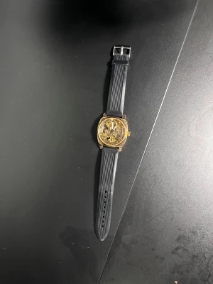 Automatisk guldig klocka med svart band - Säljer en automatisk klocka med transparent urtavla där du ser hela det mekaniska verket. Klockan har en fyrkantig guldig boett med snygga detaljer och ett svart gummiband. Baksidan är också transparent och visar urverket. Perfekt för dig som gillar coola accessoarer.