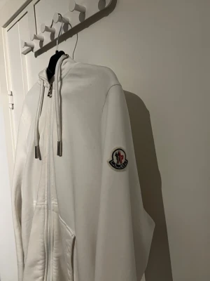 Vit hoodie från Moncler med dragkedja - Snygg vit hoodie från Moncler med hel dragkedja, huva och snörning. Tröjan har Moncler-logga på ärmen och coola ränder i rött, blått och vitt på muddar och huva. Mjuk bomullskänsla och två fickor framtill. Perfekt för dig som gillar stilrena streetwear-vibbar.