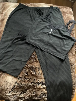 Svart ribbad pyjamas set med spets - Svart ribbad pyjamas set med smala axelband och dekorativ spets längs v-ringningen. Toppen har två små knappar framtill och är croppad i modellen. Och byxorna i rak model med snörning fram.  Materialet är mjukt och stretchigt, perfekt för chill eller layering. Snygg detalj med spetsen som ger en extra touch.