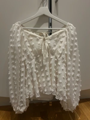 Vit blus med puffärm och knyt - Superfin vit blus med puffiga långärmar och coola 3D-prickar över hela tyget. Blusen har en söt knytning fram och är gjord i ett lätt, transparent material med ett innerlinne. Perfekt för dig som gillar romantiska och trendiga detaljer.