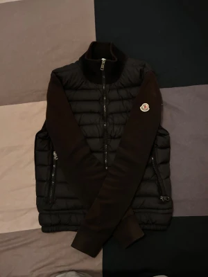 Moncler cardigan - Svart pufferjacka från Moncler med quiltad front och stickade ärmar. Jackan har två sidofickor med dragkedja, hög krage och Moncler-logga på vänster ärm. Materialmix av polyester och stickad textil ger en clean och modern look. Lite solbränd