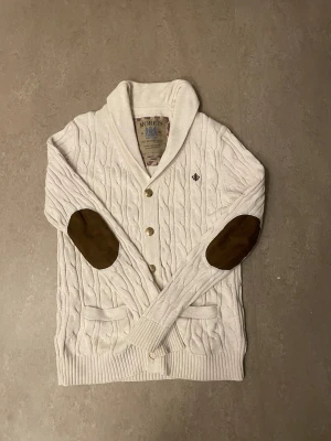 Kabelstickad morris cardigan - Vit/beige kabelstickad kofta från Morris med bruna mockadetaljer på armbågarna, två fickor framtill. Snygg krage och broderad logga på bröstet ger en klassisk och chill vibe. Perfekt för lager-på-lager under höst och vinter.