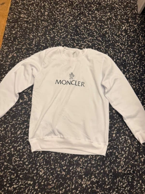 Vit sweatshirt från Moncler, storlek L - Vit sweatshirt från Moncler i storlek L med klassisk rund hals och svart Moncler-logga tryckt på bröstet. Tröjan har långa ärmar och ribbade muddar vid hals, ärmslut och nederkant. Perfekt för dig som gillar stilrena och exklusiva märken. Passar också storlek M