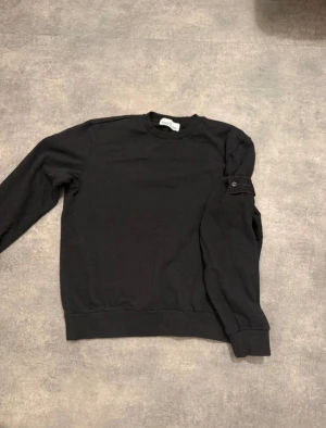 Svart sweatshirt från Stone Island - Svart sweatshirt från Stone Island med klassisk rund hals och långärmad design. Tröjan har en ikonisk patch på vänster ärm och är tillverkad i mjuk bomull. Passformen är normal och ribbade muddar finns vid ärmslut och nederkant. Perfekt för en clean och stilren look.