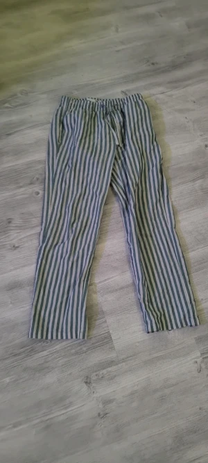 Randiga linnebyxor med snörning - Säljer ett par chill randiga linnebyxor med gröna, blå och vita ränder. Byxorna har elastisk midja med snörning och raka ben för en avslappnad vibe. Perfekta för varma dagar eller softa hemma. Superlätta och luftiga!