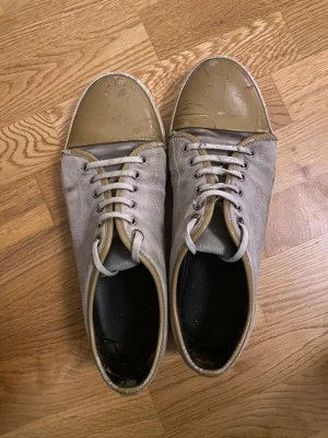 Lanvin sneakers med grå mocka och beige tå - Lanvin sneakers med grå mocka och beige lackad tå. Klassisk rund tå och platt sula, med vita snören och metallöglor. Snygg kontrast mellan material och färg, perfekt för dig som gillar stilrena och unika sneakers.