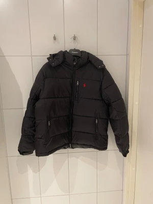 Polo Ralph Lauren Pufferjacka – Svart - Säljer en svart Polo Ralph Lauren-jacka som inte passar min stil. Märkt XL, men sitter mer som L/M.  Nypris 4 500–5 000 kr. Skick: Fint begagnat. Äkthet: QR-koden fungerar och leder till den officiella hemsidan. Detaljer: Avtagbar huva, två fickor med dragkedja, varm och lätt.  Mått: 	•	Längd: 64 cm 	•	Axelbredd: 50 cm 	•	Ärmar: 60–63 cm 