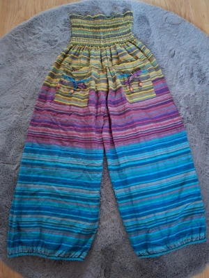 Färgglada randiga bomullsbyxor - Säljer ett par sjukt sköna och färgglada byxor i 100% bomull, tillverkade i Indien. Byxorna har hög smockad midja, breda ben och elastiska benslut. Mönstret är randigt i blått, lila, rosa, gult och grönt. Framtill finns två fickor med snörning och pärlor.