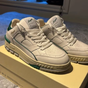 Axel Arigato vita sneakers med gröna detaljer - Säljer ett par fräscha vita sneakers från Axel Arigato med gröna detaljer och logga på sidan. Skorna har snörning, perforerade partier och en chunky sula med gröna inslag undertill. Tillverkade i skinn för en lyxig känsla och stilren look. Perfekta för dig som gillar streetwear. Skorna är helt oanvända och säljer dem pågrund av att de aldrig kommit till användning 