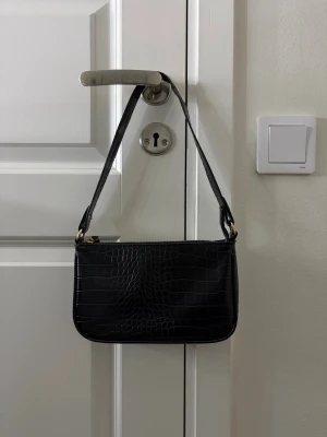Svart axelväska i crocopräglat skinn - Snygg svart axelväska med crocopräglat mönster och guldfärgad dragkedja. Väskan har en stilren, rektangulär form och ett kort axelband. Perfekt för dig som gillar minimalistisk och trendig design.