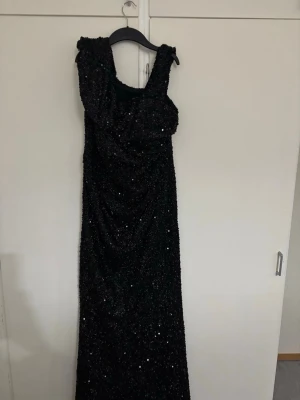 Svart glittrig långklänning med paljetter - Säljer en svart långklänning med glittrande paljetter över hela klänningen. Klänningen har breda axelband och en asymmetrisk detalj vid axeln som ger en cool och unik look. Perfekt för dig som vill sticka ut med en glamorös stil.