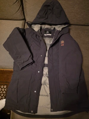 Svart dunjacka Henri Lloyd XL - Säljer en svart dunjacka från Henri Lloyd i storlek XL. Jackan har huva, stora fickor och snygga broderade detaljer med logga och brittisk flagga på bröstet. Insidan är quiltad i grått och jackan stängs med både dragkedja och knappar. Perfekt för kalla dagar.