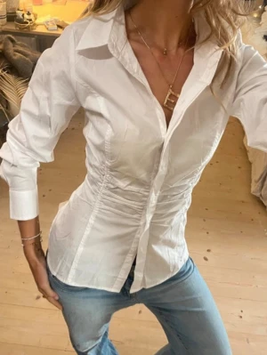 Vit figursydd skjorta med knappar - Säljer en vit skjorta med figursydd passform och snygga rynkade detaljer vid midjan. Köpt på vinted för 800kr men var tyvärr för liten för mig