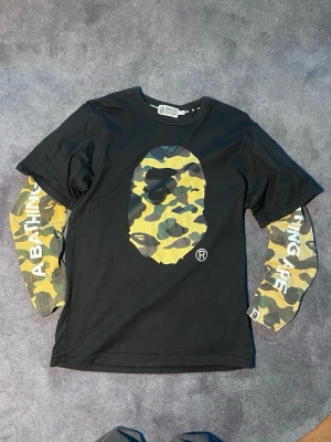 Svart BAPE t-shirt med camo tryck - Säljer en svart t-shirt från A Bathing Ape i storlek M. T-shirten har ett stort camo-tryck med BAPE-loggan på bröstet och långa ärmar i klassiskt grönt camouflagemönster med vit text. Perfekt för dig som gillar streetwear och vill sticka ut.