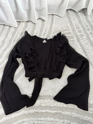 Svart omlottblus med volanger från NA-KD - Säljer en svart omlottblus från NA-KD med långa, vida ärmar och snygga volangdetaljer framtill. Blusen har knytband i midjan och är tillverkad i ett lätt, transparent tyg med diskret prickstruktur. Perfekt för dig som gillar trendiga och feminina plagg.