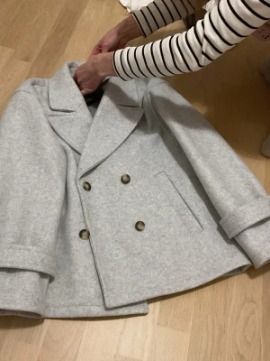 Ljusgrå skepparkavaj från RYVLS - Säljer en stilren ljusgrå skepparkavaj från RYVLS med dubbelknäppning och breda slag. Jackan har långa ärmar, två sidofickor och snygga knappar framtill. Perfekt för dig som gillar klassisk och clean look.