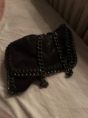 Svart axelväska med kedjedetaljer - Säljer en svart axelväska med glansig yta och grova kedjor längs kanterna och som axelrem. Väskan har en mjuk form och är tillverkad i mocka med metallkedjor som ger en cool och edgy vibe. Perfekt för dig som gillar statement-accessoarer.