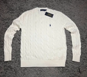 Vit stickad tröja Polo Ralph Lauren - Vit kabelstickad tröja från Polo Ralph Lauren med klassisk blå broderad logga på bröstet. Tröjan har rund hals, ribbade muddar och är långärmad. Perfekt för dig som gillar clean och tidlös stil.ett till ett. Fraktar snabbt:)