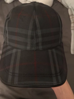  Burbery - Snygg svart Burbery keps med diskret rutigt mönster i grått och röda linjer. Klassisk form med böjd skärm och justerbar baksida. Perfekt för dig som vill ha en stilren accessoar med lite edge.