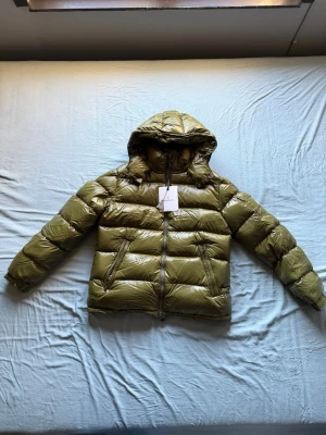 Moncler Maya - Säljer en olivgrön pufferjacka från Moncler med glansig finish och huva. Jackan har två snedställda fickor med dragkedja, tydlig Moncler-logga på ärmen och kraftig dragkedja framtill. Perfekt för kalla dagar och riktigt snygg streetstil. Helt oanvänd