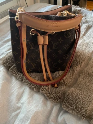 Väska - Säljer en snygg med klassiskt monogrammönster i mörkbrunt och beige. Väskan har guldfärgade metalldetaljer, justerbar axelrem och snörning upptill. Materialet är skinn och den har en rymlig bucket-form som är både trendig och praktisk.