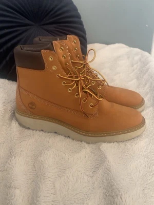 Timberland boots i brun skinn, strl 41 - Säljer ett par klassiska Timberland boots i brun skinn med ljus sula och gul snörning. Skorna har rund tå, högt skaft och vadderad krage i mörkbrunt. Perfekta för dig som gillar street och outdoor vibes.