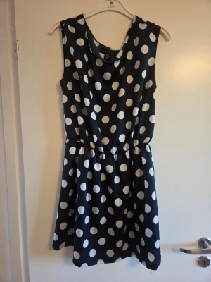 Svart prickig klänning från Amisu, stl 38 - Snygg svart kortklänning från Amisu med stora vita prickar. Klänningen är ärmlös och har en rund halsringning samt ett lätt rynkat midjeparti för en skön passform. Perfekt för dig som gillar retrostil och vill sticka ut med ett coolt mönster.