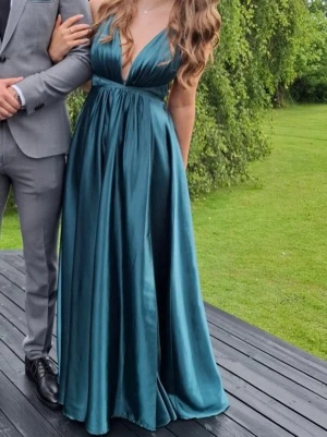 Mörkblå långklänning i satin med öppen rygg - Säljer nu min älskade balklänning, en elegant mörkgrön långklänning i satin med djup v-ringning och tunna axelband. Klänningen har öppen rygg med smala band och ett vackert fall med en gömd slits. Perfekt för bal eller andra speciella tillfällen. Det har även sytts fast ett litet spänne för att kunna täcka den nedersta delen av v-ringningen, samt sytt upp den lite. Jag är 1,64 passar perfekt. Använts en gång.