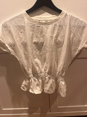 Vit blus med volang från H&M - Säljer en vit blus från H&M med rund hals och korta, vida ärmar. Blusen har små pärldetaljer på framsidan och en bred volang nedtill med smockad midja. Supersöt! 