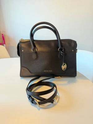  Michael Kors Harrison Black  - Stilren svart handväska från Michael Kors i skinn med guldfärgade detaljer och logotyp-charm. Väskan har både handtag och avtagbar axelrem samt dragkedja upptill. Klassisk och elegant design som funkar till allt.