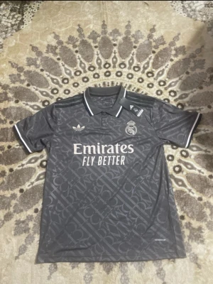 Grå Real Madrid x Adidas tröja - Säljer en grå Real Madrid x Adidas tröja med mönstrad design och vita detaljer. Tröjan har krage med vit kant, korta ärmar och tryck med 'Emirates Fly Better' på bröstet. Perfekt för dig som gillar fotboll och snygga sportkläder.