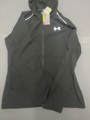 Svart träningsjacka Under Armour - Svart träningsjacka från Under Armour med huva och dragkedja. Jackan har vita detaljer vid axlarna och en stor logga på ryggen. Materialet är lätt och andas, perfekt för löpning eller gym. Regular fit och tillverkad i syntet för att hålla dig torr och bekväm. Storlek L men den sitter lite tajtare än vanligt så jag tror att den skulle passa M