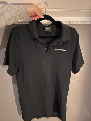Svart Peak Performance piké S - Svart pikétröja från Peak Performance i storlek S. Klassisk krage och knappslå framtill, med diskret logga broderad på bröstet. Perfekt för en clean och sportig look. Materialet känns som bomull och är mjukt mot huden.