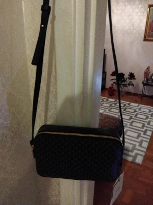 Svart axelväska med mönster - Snygg svart axelväska med geometriskt mönster och guldfärgad dragkedja. Väskan har en justerbar axelrem och rektangulär form, perfekt för att bära dina viktigaste grejer. Materialet ser ut att vara syntet och designen är stilren med diskreta detaljer.