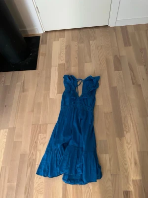 Blå långklänning med volang - Säljer en blå långklänning med v-ringad hals och volangdetaljer upptill. Klänningen har knyt i ryggen och är ärmlös, med en luftig och flowy passform. Perfekt för varma dagar och har en bohemisk känsla.