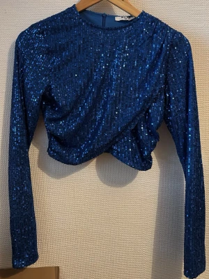 Blå paljettopp från NA-KD - Säljer en glittrig blå topp från NA-KD med paljetter över hela ytan. Modellen är croppad med lång ärm och har en rund halsringning samt dragkedja i ryggen. Perfekt för dig som vill sticka ut med en festlig look. Toppens material är polyester.