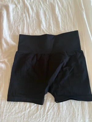 Svarta ribbade träningsshorts H&M - Svarta träningsshorts från H&M Sport med hög midja och ribbad struktur. Stretchigt syntetmaterial som sitter skönt och följer kroppen. Perfekta för gymmet eller löprundan. Snygg, enkel design med diskreta vertikala ränder. Aldrig använda då de var för små.
