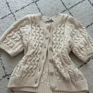 Beige kabelstickad kofta från Zara - Superfin beige kabelstickad kofta från Zara med stora matchande knappar och kortare ärmar. Koftan har ett chunky stickat mönster och rund halsringning, perfekt att slänga över en topp för en mysig look.(säljer den för att den är för stor