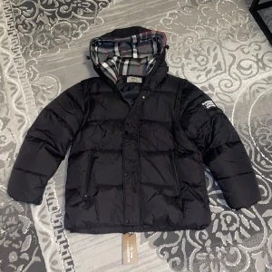 Svart pufferjacka från Burberry - Svart pufferjacka från Burberry med klassisk rutig huva i grått, vitt och rött. Jackan har dragkedja, knappar och två fickor framtill. Märkespatch på ärmen och snyggt quiltad design. Perfekt för kalla dagar och riktigt stilren look.