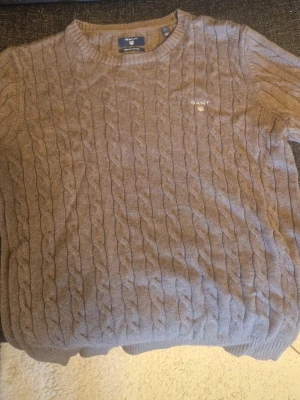 Brun stickad tröja från GANT - Säljer en brun stickad tröja från GANT med klassiskt kabelstickat mönster. Tröjan har rund halsringning och ribbade muddar vid ärmslut och nederkant. Perfekt för dig som gillar stilrena och tidlösa plagg.