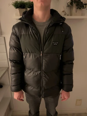 Svart pufferjacka från Alessandro - Säljer en svart pufferjacka från Alessandro med mörkgrå panel upptill och huva med snörning. Jackan har dragkedja framtill, två sidofickor med dragkedja och en snygg logga på bröstet. Perfekt för kalla dagar och riktigt skön att bära.