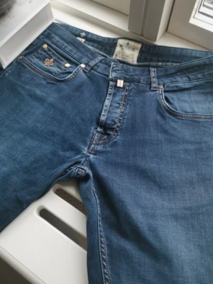 Blå jeans från Morris med kontrastsömmar - Snygga blå jeans från Morris med klassisk femficksdesign och kontrastsömmar. Jeansen har en mörkblå ton, bruna detaljer på fickorna och ett läderpatch med Morris-logga bak. Materialet är slitstarkt jeans och passformen är normal.