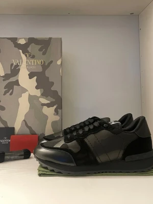 Valentino Rockrunner  - Svarta Valentino Rockrunner sneakers med coolt kamouflagemönster och detaljer i mocka och skinn. Snygg platt sula och rund tå, klassisk snörning och exklusiv känsla. Perfekt för dig som vill sticka ut med ett lyxigt streetwear vibe.