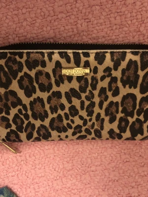 Leopardmönstrad plånbok Don Donna - Säljer en snygg leopardmönstrad plånbok från Don Donna med guldfärgade detaljer och dragkedja. Plånboken har flera fack för kort och mynt, och är tillverkad i syntetmaterial med beige och svarta toner. Perfekt för dig som gillar djurmönster och vill ha ordning på dina grejer.